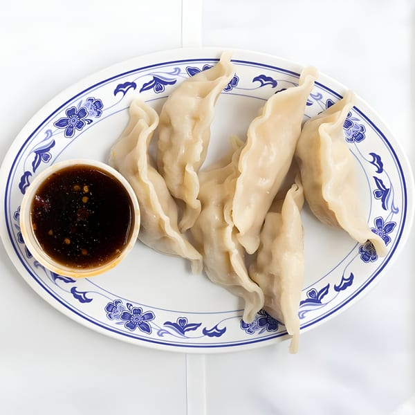 A white oval plate with blue floral patterns holds six dumplings and a side of dark dipping sauce.